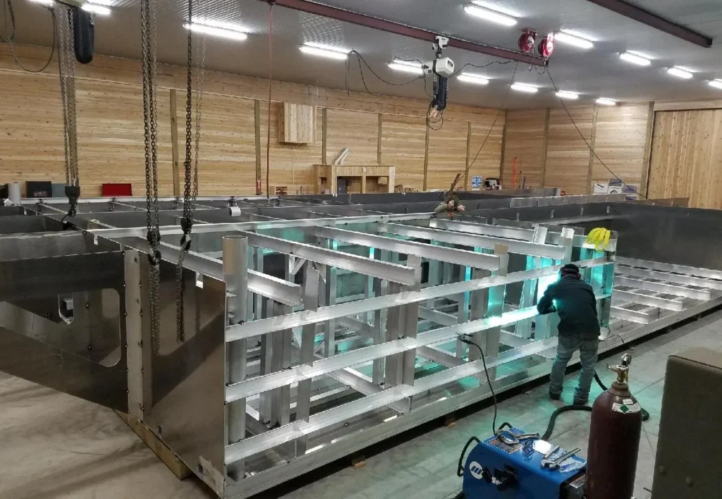 Warehouse with wood plank walls, overhead florescent lights, large aluminum frame under construction with man arc welding on it